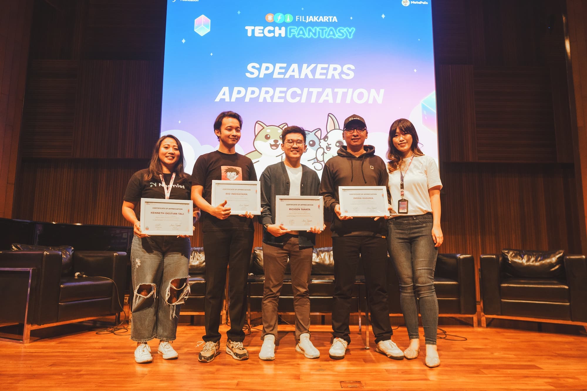 Group of five people standing on stage holding certificates of appreciation with a "Tech Fantasy" event banner in the background